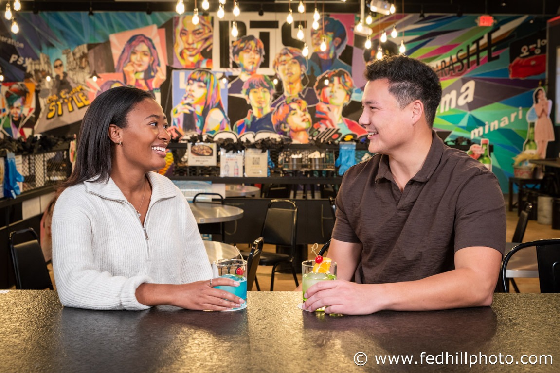 Tourism/lifestyle/restaurant photo of two people sitting at a bar holding drinks and smiling at each other.