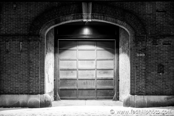 Fine art black and white photograph of footprints in snow past a fire station at night in Baltimore City, Maryland.