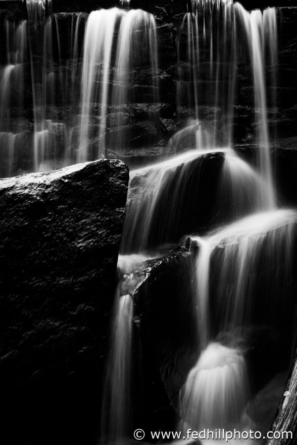 Black and white fine art photo of waterfall over stone dam, splashing on rocks.